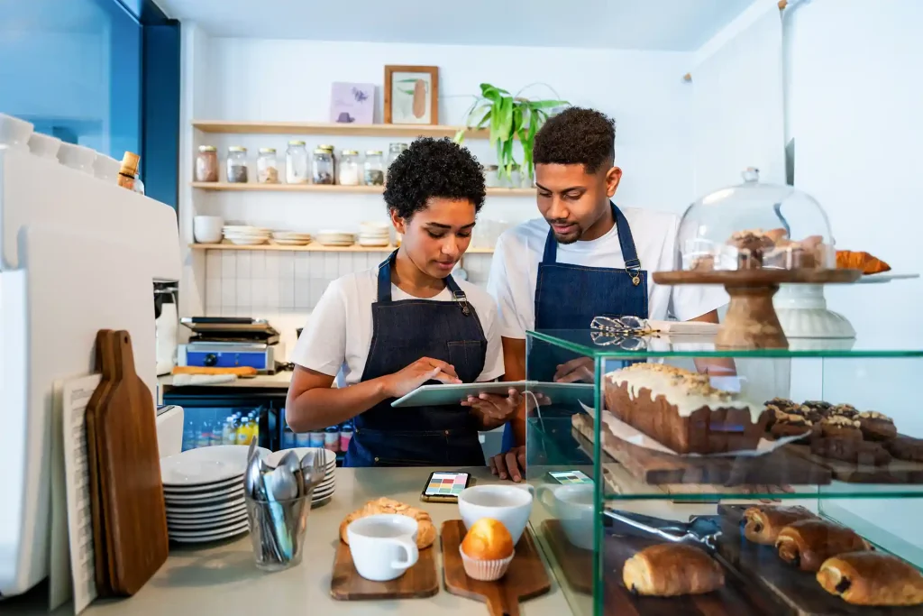 En man och en kvinna står bakom en bardisk i ett kafé och kollar på en surfplatta. På bordet ligger en telefon med Onslip kassasystem på skärmen. Till höger om bilden framför mannen och kvinnan ser man bakelser och bröd. Till vänster ser man en kaffemaskin och muggar.