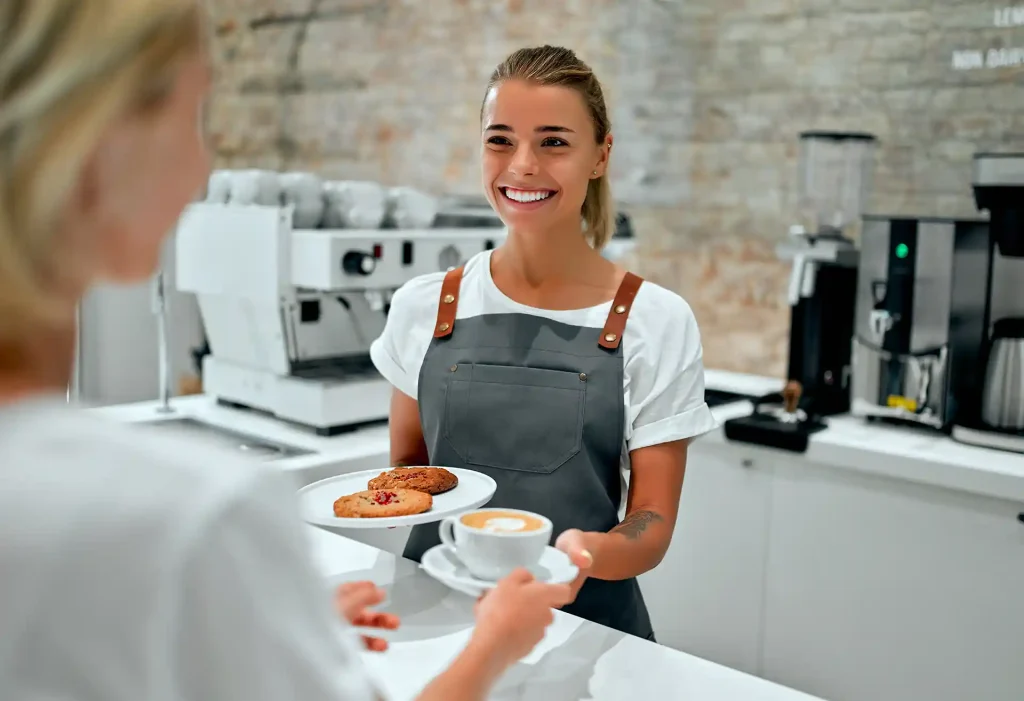 En kvinna som står bakom en bardisk på ett kafé och har fikabröd i ena handen och i den andra en kopp kaffe. Kvinnan serverar en gäst som tar emot det kvinnan har i händerna.