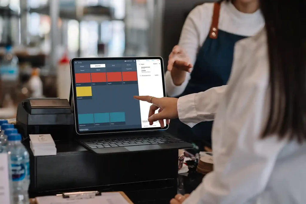 En person slår in produkter på ett kassasystem på en surfplatta. Bakom surfplattan står en person med förkläde.