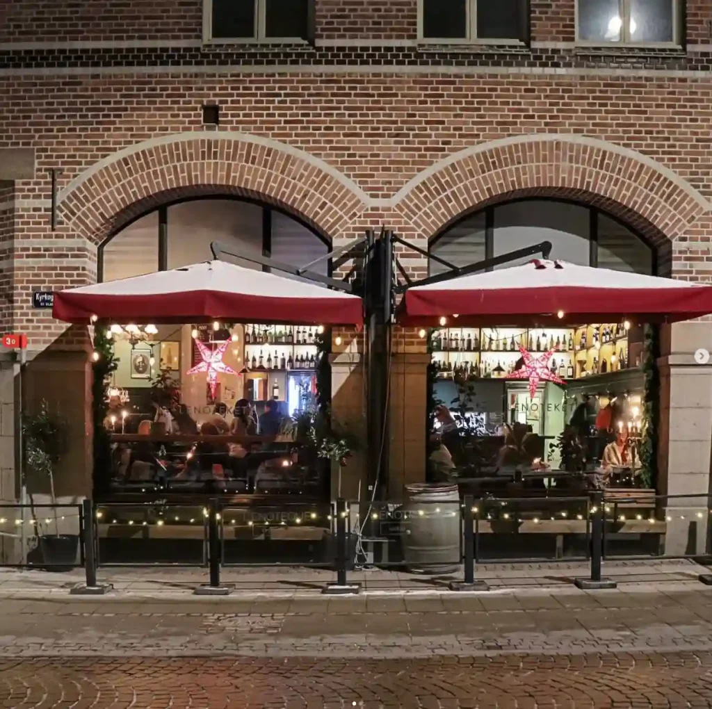 Mörk bild på en tegelbyggnad med två röda parasoller framför entrén in till en restaurang som finns i byggnaden. Genom de stora fönsterpartierna ser man in i restaurangen och folk som sitter vid bord.