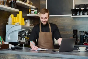 EN glad man bakom en bardisk i ett kafé som håller i en penna och skriver på ett vit skrivblock. Till höger i bilden står det en laptop framför personen på bardisken. I bakgrunden bakom personen står en silvrig kaffemaskin med gula kaffemuggar.