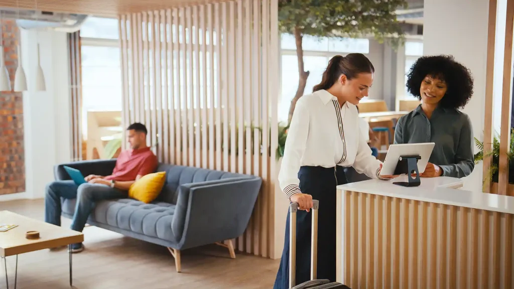 Två kvinnor står vid en surfplatta i en hotelllobby och checkar in. Den ena kvinnan som står närmast i bild håller i en resväska med hjil och har på sig en bit skjorta och har mörkt brunt håret uppsatt i hästsvans. Den andra kvinnan har stort krulligt hår med svart hårfärg. Surplattan som de kollar på står på ett stativ på en receptiondisk som har en vit stenskiva och träpaneler. I bakgrunden ser man en man sitta i en blå soffa med en dator i knät. Han sitter bredvid en gul kudde som han har nära sig. Mannen har på sig en orange tröja. Framför sovan står ett soffbord med en ljus träskiva på. Hotell lobbyn släpper in mycket dagsljus och ser trevlig ut. Många ljusa färger i inredningen.