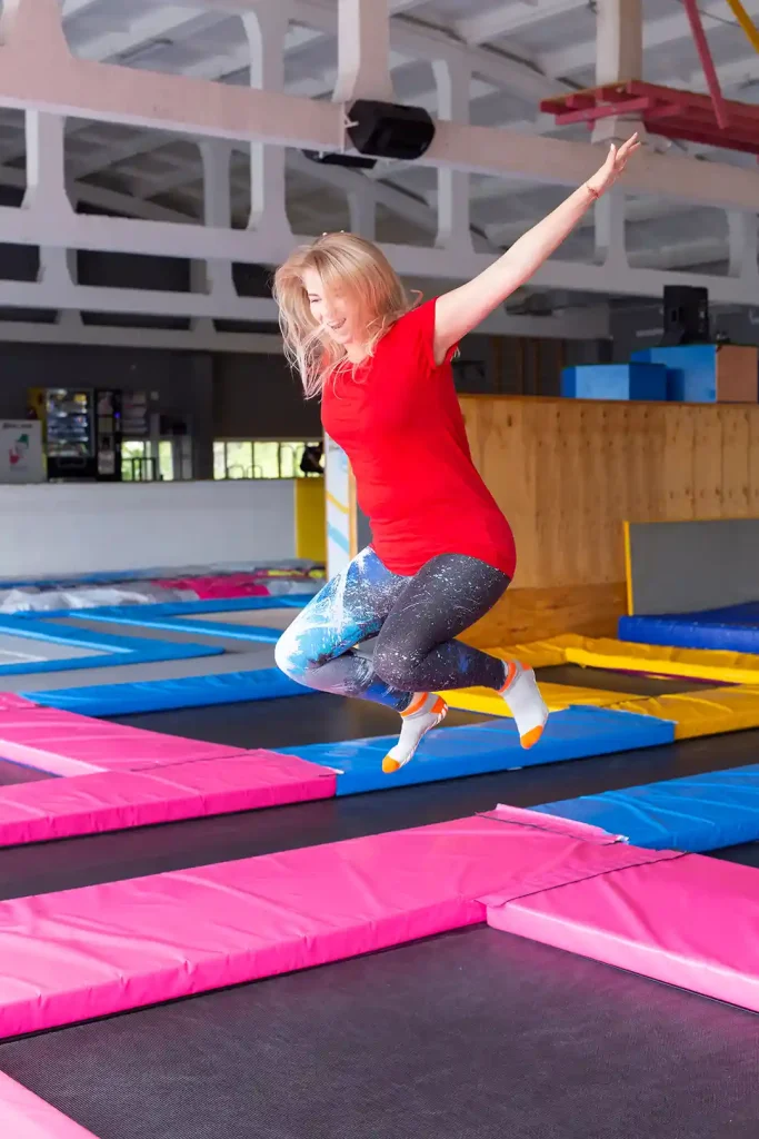 En kvinna som är i ett nöjescenter med flera trampoliner och hoppar. Kvinnan har långt blont hår och har röd tshirt och har på sig strumpor.