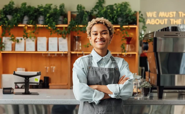 En kvinna kort krulligt ljust hår som står med armarna i kors. Kvinnan står framför en bardisk i en restaurang med ett förkläde och ljus blå skjorta på sig. Kvinnan ler stort. På bardisken i bakom till vänster i bild står en svart kortterminal och ett POS system på en surfplatta som hålls upp av ett svart metall stativ för surfplattor. Till höger i bild ser man en kaffemaskin och en några små krukväxter. I bakgrunden bakom bardisken står finns en hylla i trä och på hyllorna finns menyer, vinglas och gröna växter som slingrar sig ned från översta hyllan.