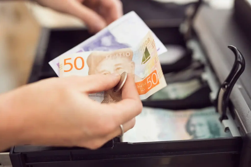 En hand som håller i svenska kontanter. En femtiolapp och en tjugolapp som är av det äldre slaget. Personerna håller händerna ovanför en kassalåda och har precis tagit ut kontanterna från kassalådan.
