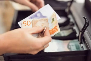 En hand som håller i svenska kontanter. En femtiolapp och en tjugolapp som är av det äldre slaget. Personerna håller händerna ovanför en kassalåda och har precis tagit ut kontanterna från kassalådan.