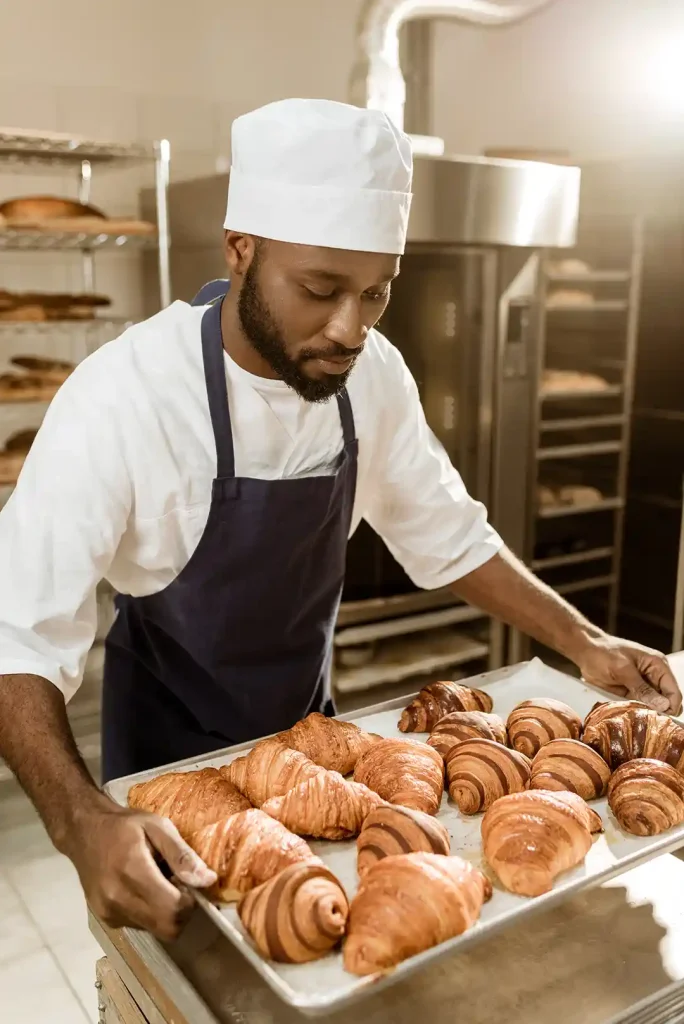 En manlig bagare med vita kläder och ett mörk förkläde lyfter en plåt med croissanter på. Bagaren står i ett bageri med en bakugn bakom sig och två vagnar med plåtar med bakverk på.