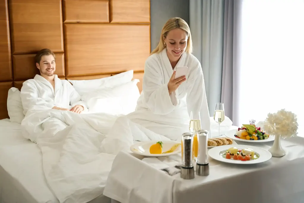En man och en kvinna sitter i sitt hotellrum med vita morgonrockar i sängen och har beställt roomservice. I sängen finns vita bäddsät och kuddar. Sängen har en brun sänggavel. Mannen har brunt kort hår och sitter lutad mot sänggaveln och ler mot kvinnan som sitter närmare i bild. Kvinnan med blont hår i bilden sitter vid slutet av sängen och har på sig en vit morgonrock. I handen håller hon en telefon och fotar av maten som de har beställt från roomservice via telefonen. På bordet som står vid sängen som kvinnan sitter när finns olika frukter och drycker.