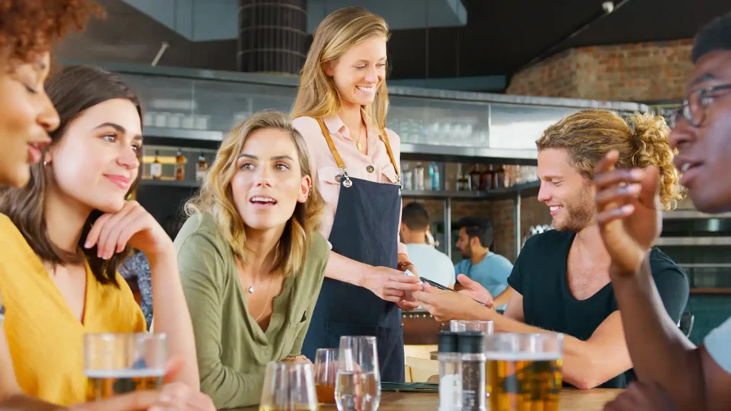 Flera personer som sitter vid ett bord i en restaurang. I bakgrunden tar en servitris betalt för den uppdelade notan av en man med långt hår. Alla personer ser glad ut och har en konversation.