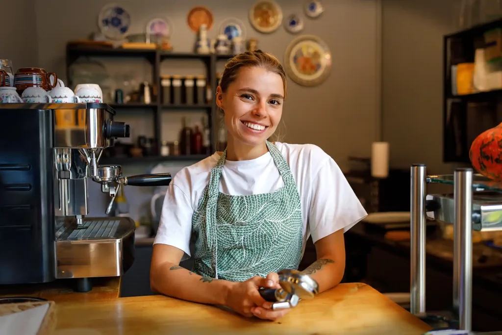 En kvinnlig barista med ljus grönt förkläde på sig som står lutad mot en bardisk och bredvid kvinnan står en kaffemaskin i café. På bardisken står även kortterminal för kortbetalningar.